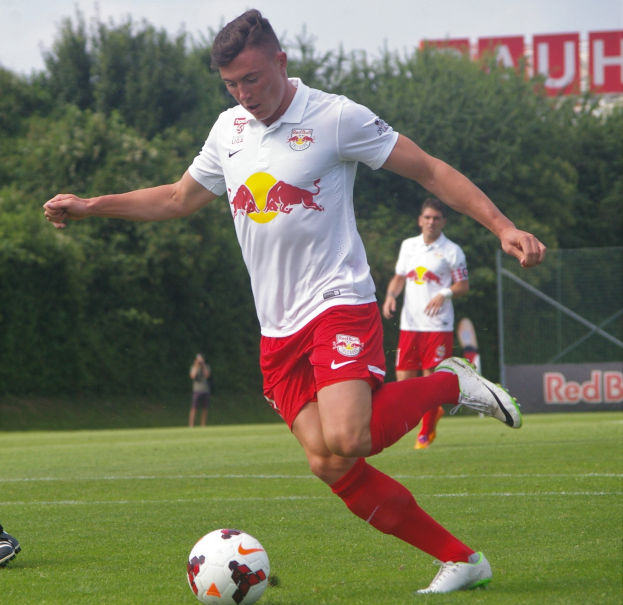 Ein Mann in einem Fußballtrikot kickt einen Ball auf einem grünen Feld mit Bäumen und einem klaren blauen Himmel im Hintergrund, mit einem Schild im Hintergrund, auf dem "RB Leipzig" steht.