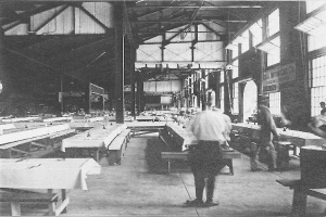 Schwarzes und weißes Foto von Menschen, die in einer Fabrik mit Maschinen arbeiten, auf dem Boden und erhöhten Flächen stehen, unter einem Dach mit Metallstreben, mit Fenstern und Säulen zu sehen, und Text am unteren Rand.