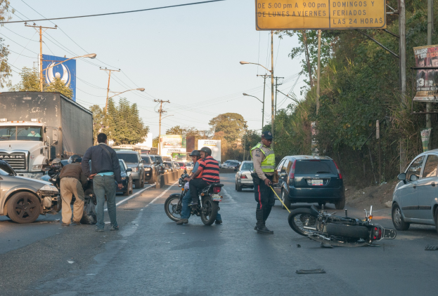 Eine Gruppe von Menschen steht neben einem verunglückten Motorrad am Straßenrand mit Fahrzeugen, Bäumen, Pfählen und Lichtern im Hintergrund.