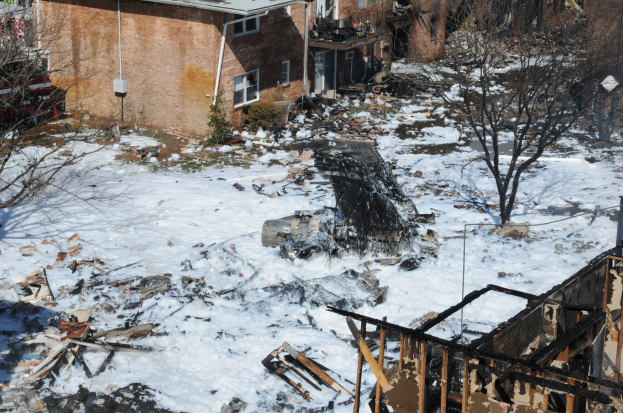 Verbrannte Überreste eines durch Feuer zerstörten Hauses in einer verschneiten Wohngegend mit Bäumen und Gebäuden im Hintergrund.