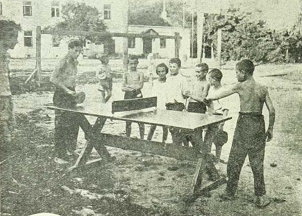 Eine Gruppe von Kindern spielt Tischtennis vor einem Gebäude mit Fenstern, umgeben von Bäumen und Pfählen, in einem Schwarz-Weiß-Bild mit Text unten.