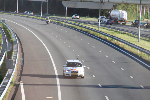Ein Auto auf einer Autobahn mit einem stationären Polizeiwagen am Rande, anderen fahrenden Fahrzeugen und Bäumen, Geländern, Pfosten und Planken im Hintergrund.