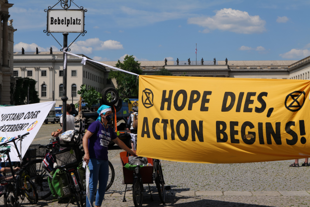 Eine Gruppe von Menschen steht vor einem Gebäude und hält ein gelbes Banner hoch, auf dem "Hope Dies, Action Begins" steht. In der Nähe parken Fahrräder und es gibt einen Pfosten mit einer daran befestigten Tafel, unter einem klaren blauen Himmel mit Bäumen im Hintergrund.
