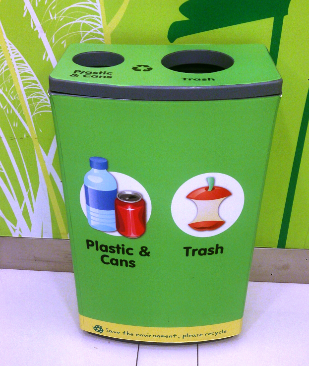 Ein grüner Recycling-Eimer voller Plastik und Dosen steht auf einem weißen Boden, mit einem Plakat im Hintergrund.