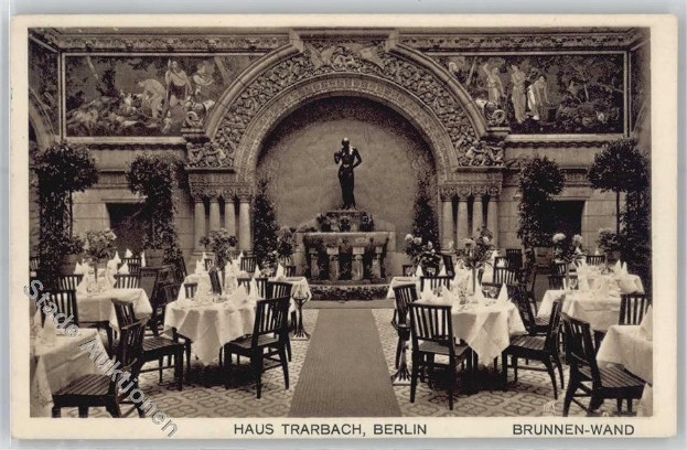Schwarz-weißes Foto eines alten Berliner Restaurant-Interieurs mit Tischen, Stühlen, Blumenvasen, einer Statue, Wandskulpturen, Pflanzen und Text unten.