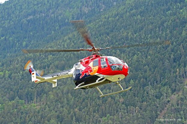 Roter Bullen-Hubschrauber über einem grünen Wald mit Bäumen auf Höhen und einem klaren blauen Himmel.
