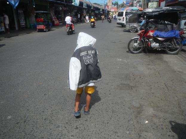 Ein junger Junge geht die Straße entlang mit Motorrädern und anderen Fahrzeugen, flankiert von Gebäuden mit Schildern, unter einem klaren blauen Himmel mit Bäumen und Pfählen im Hintergrund.