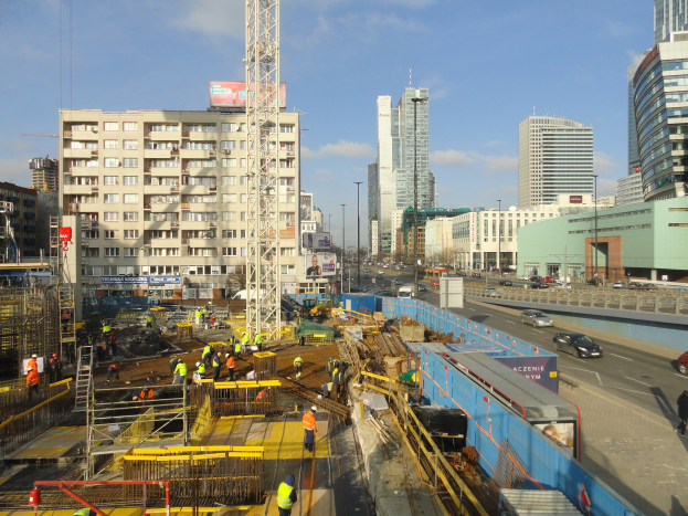 Eine Baustelle in einer Stadt mit Arbeitern, Fahrzeugen, Metallrahmen, Polen, Schildern, Gebäuden und einem bewölkten Himmel.