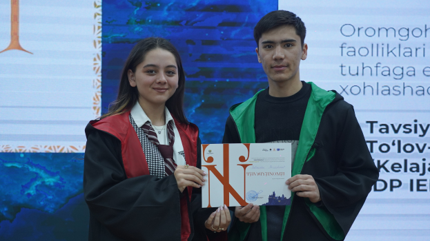 Ein Mann und eine Frau in Graduationsroben stehen Seite an Seite, jeder hält ein Zertifikat in der Hand, mit einem Bildschirm im Hintergrund.