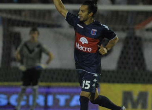 Ein Fussballspieler feiert nach einem Tor auf dem Boden, mit jubelnden Fans und einer Person im Hintergrund und einer Tafel mit Text dahinter, alles vor einem leicht unscharfen Hintergrund.