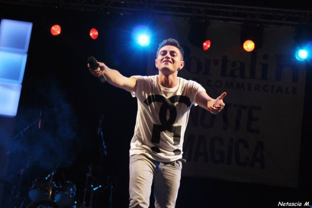 Mann auf einer hell erleuchteten Bühne mit einem Mikrofon in der Hand, im Hintergrund verschiedene Musikinstrumente und ein "Nouveau Magica"-Plakat.