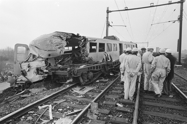 Eine Schwarz-Weiß-Szene eines Zugunglücks mit Menschen in Helmen, die herumstehen, Strommästen, Bäumen, Gebäuden und einem klaren Himmel im Hintergrund.