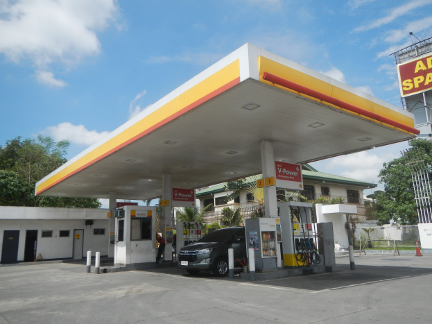 Ein Auto steht vor einer Tankstelle, mit Gebäuden, Bäumen und einem klaren blauen Himmel im Hintergrund und einer Werbetafel mit dem Wort "Petrol" auf der rechten Seite.