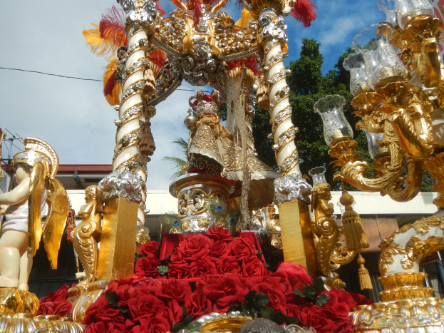 Ein Paradewagen mit roten Rosen und goldenen Verzierungen, darunter Statuen und Blumen, mit einem Gebäude, Bäumen und einem bewölkten Himmel im Hintergrund.