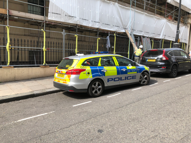 Ein Polizeiauto steht auf der Straße neben einem Gebäude, daneben stehen zwei Helme tragende Personen, ein Geländer und ein Pfahl sind im Vordergrund zu sehen, und ein weißes Tuch hängt über das Gebäude im Hintergrund, was auf eine polizeiliche Untersuchung nach einem Messerangriff in London hinweist.