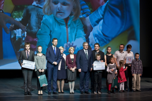 Gruppe von Menschen auf einer Bühne mit Taschen und Urkunden, im Hintergrund ein Bildschirm mit einem Bild eines Kindes und anderen