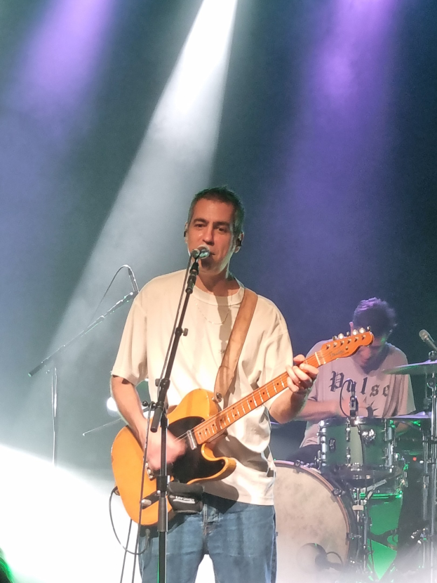Ein Mann spielt eine Gitarre und singt in ein Mikrofon auf der Bühne, während ein anderer Mann im Hintergrund Schlagzeug spielt.