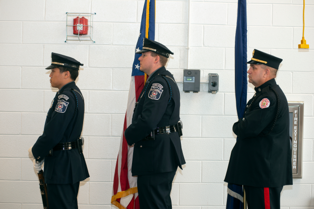 Drei Polizisten in Uniform, die Mützen tragen und Waffen halten, stehen nebeneinander mit Flaggen und anderen Gegenständen an der Wand hinter ihnen, darunter ein Feuerlöscher.
