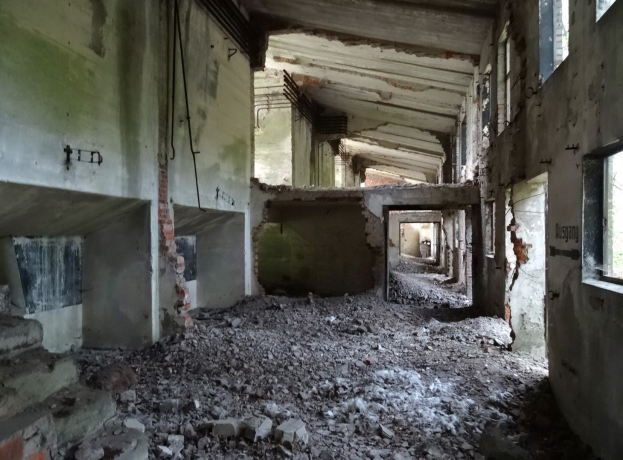 Altes Geb├Ąude in starkem Verfall mit zerbrochenen Fenstern, verstreuter Trümmers, freiliegenden Rohren und Schutt über den Boden und W├Ąnde.