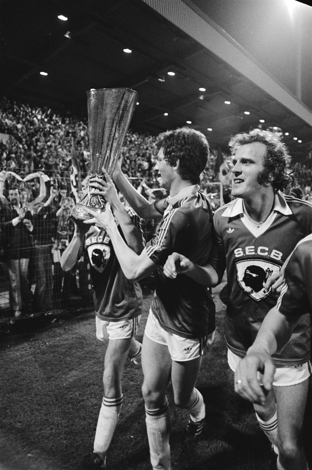 Schwarzes und weißes Foto von Fußballspielern, die einen Pokal auf einem Feld halten und ihren Sieg im FA-Cup-Finale feiern, während Zuschauer hinter einem Zaun zuschauen.