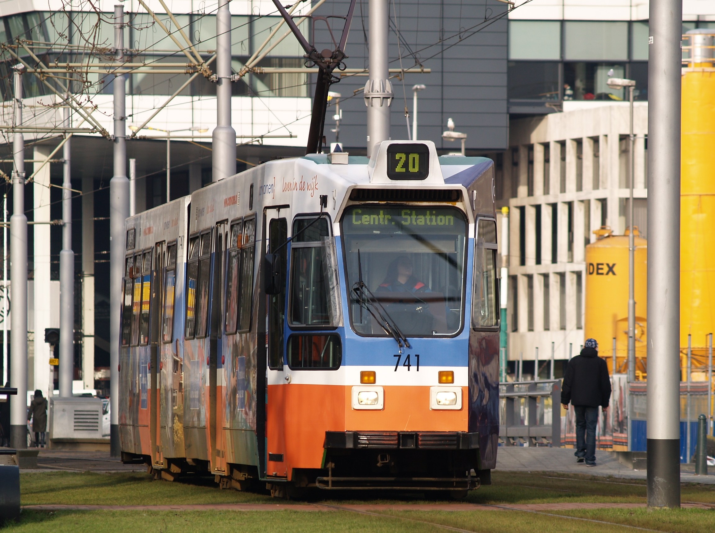 Blauer und orangefarbener Straßenbahnwagen auf einer Stadtstraße mit einem Fahrgast im Inneren, einer Person auf dem Gehweg und Gebäuden im Hintergrund.