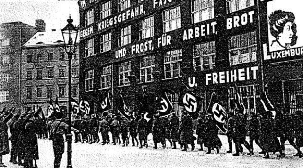 Schwarzes und wei├čes Bild einer Gruppe, die mit Fahnen und Transparenten die Stra├če entlangmarschiert, mit einem Lichtmast links und einem Nazi-Parteischild im Hintergrund.