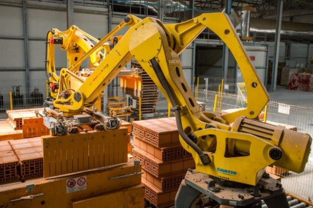 Ein großes Lagerhaus voller numerous boxes, Kräne und verschiedene industrielle Ausrüstung, mit einem Zaun und Masten im Vordergrund und einer Wand im Hintergrund.