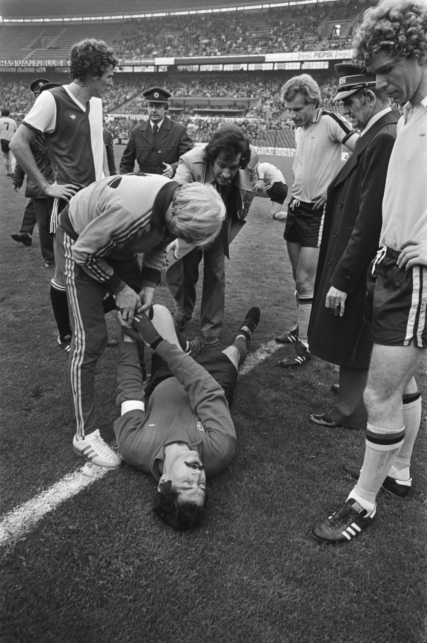 Schwarzes und weißes Bild eines Fußballspielers auf dem Boden umgeben von einer Gruppe von Männern, mit einer Menge im Stadionhintergrund; der Spieler scheint verletzt zu sein.
