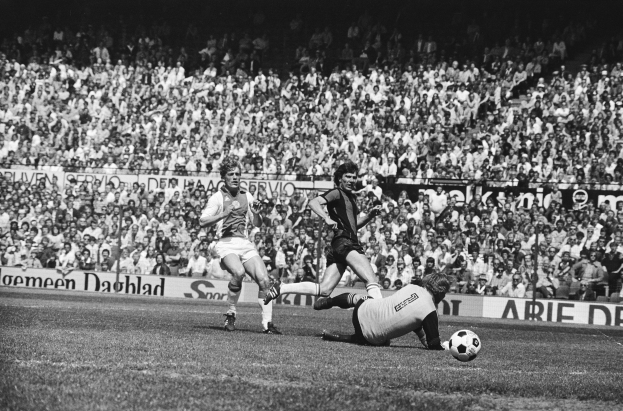 Schwarzes Foto von Fußballspielern im Einsatz während eines Bundesligaspiels zwischen Ajax und Hannover 96 in der Saison 1978-79, mit Zuschauern und einer Ligabanner im Hintergrund.