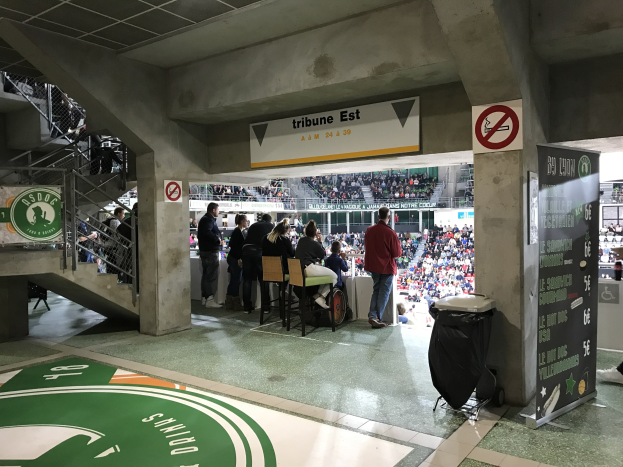 Eine Gruppe von Menschen steht vor einem sitzenden Publikum in einem Stadion, mit Schildern, die "Boston Celtics vs New York Jets", ein Müllcontainer, eine Tafel mit Text, eine Treppe mit Geländern und eine Decke mit Deckenleuchten zeigen.