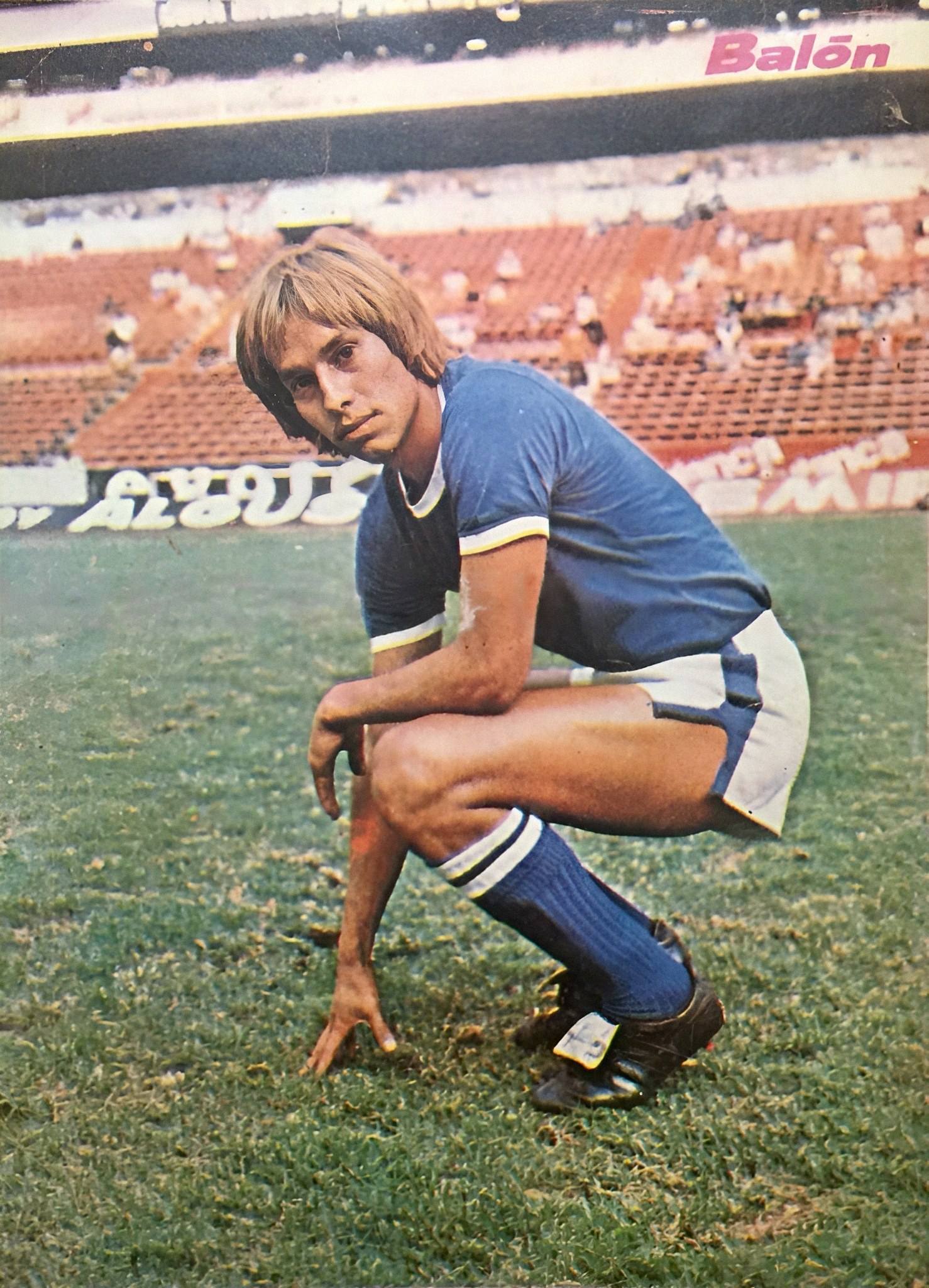 Ein Mann kniend auf einem Fußballfeld umgeben von Menschen auf Stühlen und Tafeln mit Text.