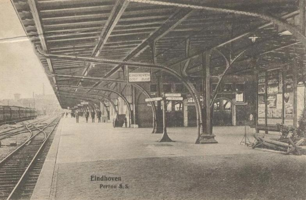 Schwarzes und weißes Foto eines Bahnhofs mit Menschen auf dem Bahnsteig, Eisenbahnschienen, einem Zug, Gebäuden im Hintergrund und dem Text "Eindhoven Post Office, Berlin, Germany" unten.