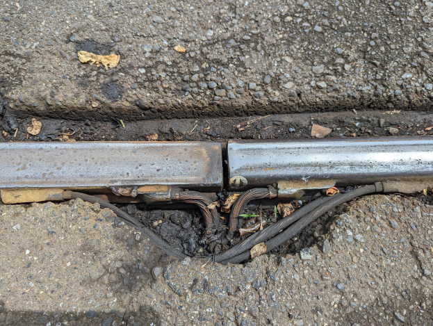 Nahaufnahme eines Metallkanalrohrs auf der Straße mit angebrachten Drähten und verstreuten trockenen Blättern.