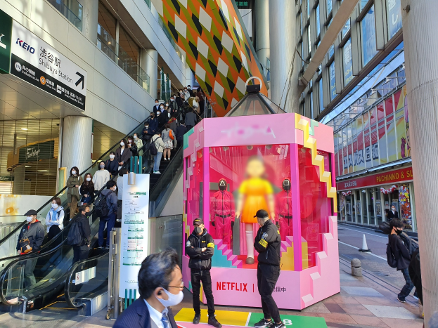 Eine Gruppe von Menschen steht vor einem Gebäude mit Glaswänden, Texttafeln, einige tragen Masken, in der Nähe einer Rolltreppe; ein pinker Kasten in der Mitte zeigt Schaufensterpuppen und trägt 'Netflix's new Netflix booth in Tokyo'.