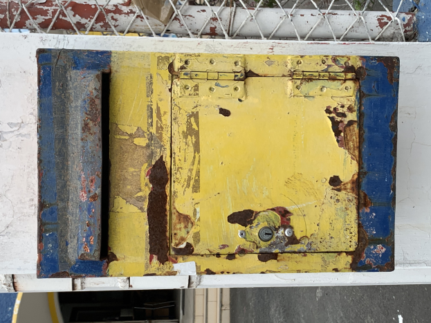 Ein gelb-blauer Metalltresor steht auf einem weißen Wall, im Hintergrund ist ein Metallzaun zu sehen.