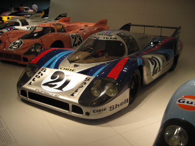 Ein Porsche 917K Rennwagen aus Le Mans in einem Museum neben anderen Sportwagen ausgestellt, mit Menschen, die zuschauen, und einem Textfeld auf der linken Seite.