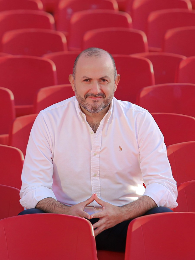 Ein Mann in einem weißen Hemd sitzt entspannt und selbstbewusst in einem roten Stadionstuhl, die Hände verschränkt, mit einem ernsten Gesichtsausdruck, während Sonnenlicht im Hintergrund scheint.