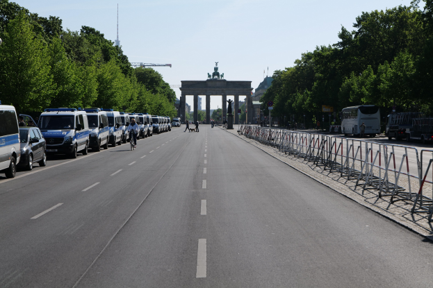 Eine Reihe von Polizeiwagen steht auf der Seite einer Straße vor dem Brandenburger Tor in Berlin, Deutschland, mit Menschen, die Fahrräder fahren und auf der Straße stehen, Absperrungen und Bäume an den Seiten und einem Bogen mit Statuen im Hintergrund.