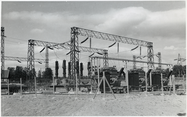 Ein Schwarz-Weiß-Foto einer elektrischen Umspannstation mit Transformatoren, Masten, Drähten, Maschinen, einem Zaun, Sand, Bäumen und einem wolkenverhangenen Himmel mit Text unten.