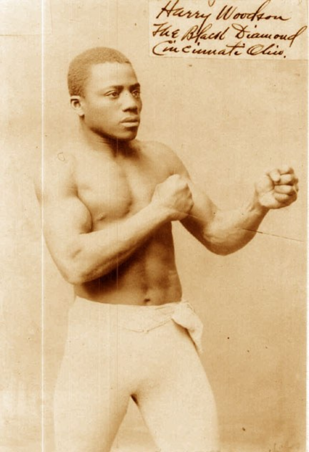 Ein Schwarz-Weiß-Foto eines Mannes in einem Boxring mit Handschuhen, Shorts und entschlossener Miene.