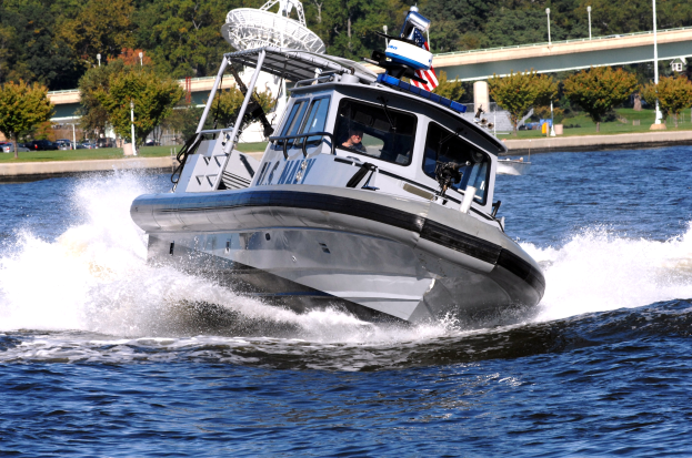 Ein Polizeiboot fährt über das Wasser mit einer Person darin, mit einer Brücke, Bäumen, Polen, Fahrzeugen, Gras und einem klaren blauen Himmel im Hintergrund.