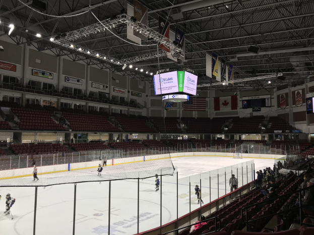 Eishockeyspiel in einer großen Arena mit einem zentralen Tor, Zuschauern auf beiden Seiten, Deckenbeleuchtung und Trägern sowie Wandbannern mit der Aufschrift "St. Louis Blues Arena."