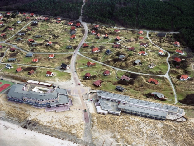 Luftaufnahme eines kleinen Strandorts mit Häusern, Straßen, Gras, Bäumen und Sand am Boden.