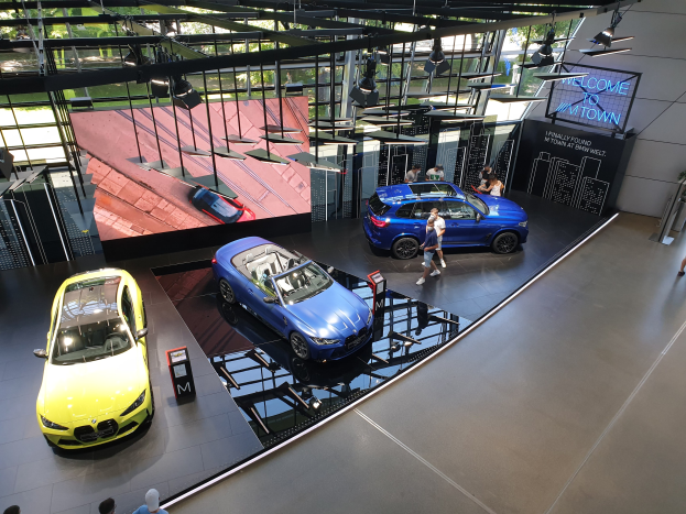 Eine Gruppe von Menschen, die im BMW Museum in München stehen, mit Autos zur Linken und einem Schild mit der Aufschrift "BMW Museum München" zur Rechten, mit Blick auf Bäume, Gebäude und den Himmel durch Glasfenster.