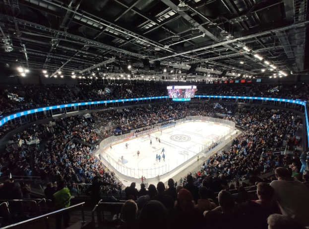 Ein Eishockeyspiel in einer großen Halle mit Zuschauern auf den Tribünen und einigen Menschen am Boden, mit Geländern, Deckenbeleuchtung und einem Bildschirm oben.