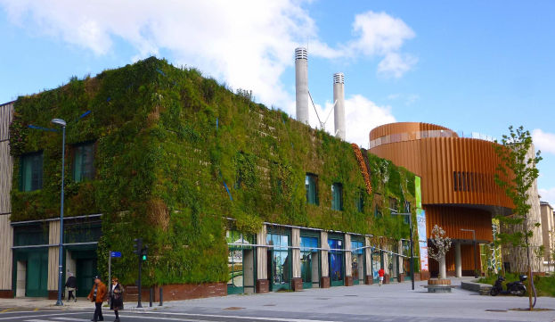 Ein großes Gebäude mit einer grünen Wand an der Seite, umgeben von Straßeninfrastruktur, Fahrzeugen, Fußgängern, Bäumen und einem bewölkten Himmel als Teil des Green Building Projects.