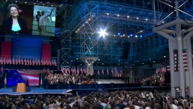 Eine große Menschenmenge sitzt vor einer Bühne mit einem Rednerpult, Mikrofon, Flaggen, einem Bildschirm und Beleuchtungsausrüstung und wartet auf eine Debatte.