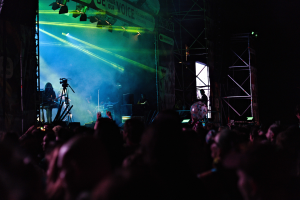Eine Menge schaut Performer auf einer hell erleuchteten Bühne bei einem Konzert zu, mit Instrumenten, Mikrofonen und einem Banner im Bild.