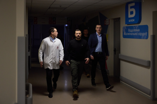 Eine Gruppe von Männern, die einen Krankenhausflur entlanggehen, mit Deckenbeleuchtung, einer Tür an der rechten Wand und Texttafeln an der linken Wand, die besorgt aussehen.