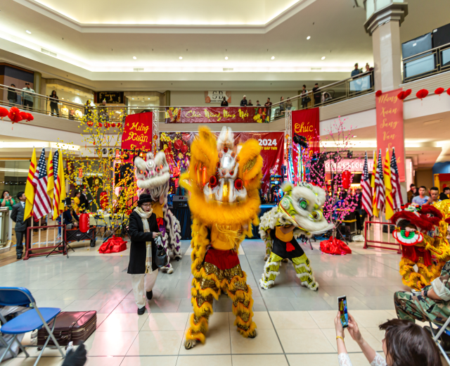 Ein lebhaftes chinesisches Neujahrsfest in einem Einkaufszentrum mit Menschen in Kostümen, die tanzen, und anderen, die auf Stühlen sitzen und mobile Telefone halten, umgeben von Fahnen, Bannern, Dekorationen und architektonischen Elementen, mit einem Wasserzeichen unten.